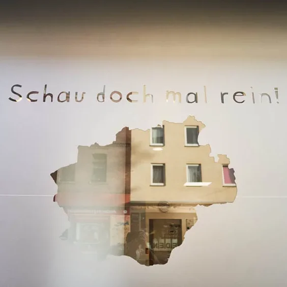 Schaufenster mit Text "Schau doch mal rein!" und Loch, das ein Gebäude zeigt.