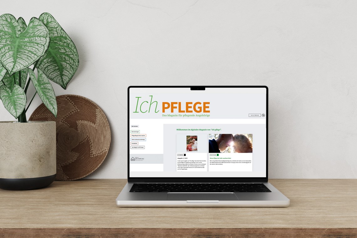 Laptop auf Tisch mit Offenem Magazin "Ich Pflege", Pflanze und Korb als Dekoration.