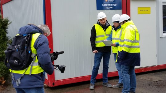 Vier Personen mit Schutzhelmen und Warnwesten vor einem Baucontainer, einer bedient eine Kamera, die anderen führen ein In