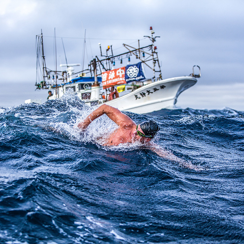 Schwimmer Andre Wiersig in stürmischem Ozean, im Hintergrund ist ein Boot zu sehen.
