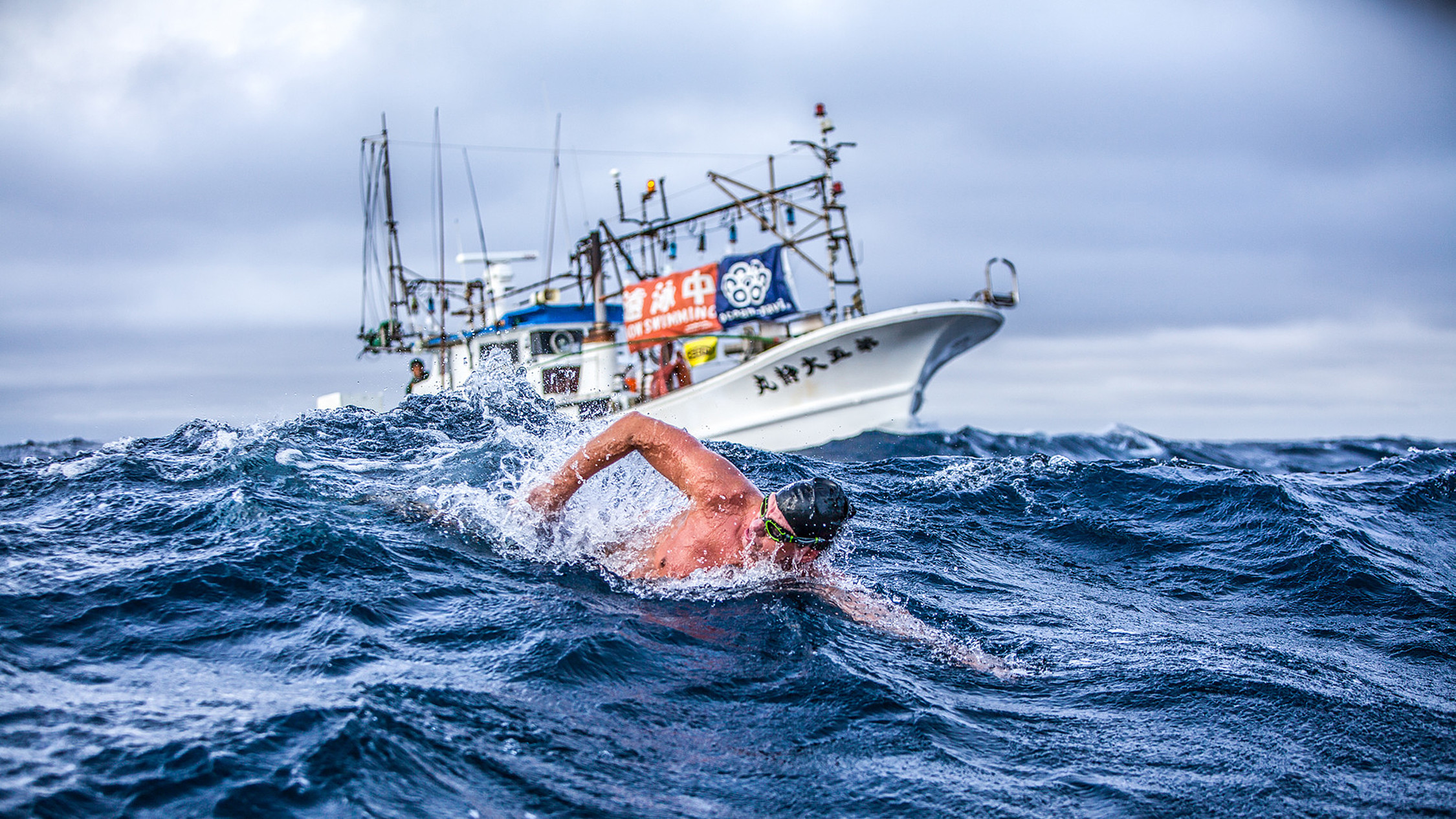 Schwimmer Andre Wiersig in stürmischem Ozean, im Hintergrund ist ein Boot zu sehen.
