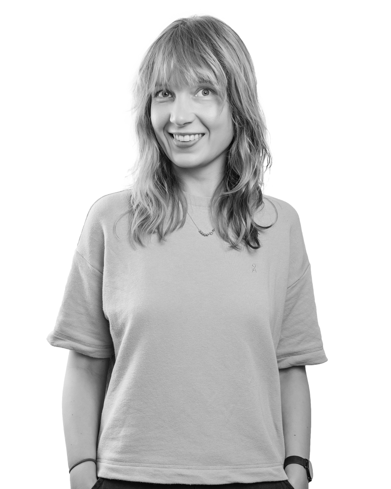 Lächelnde Frau in Flieder-Shirt mit Händen in den Taschen.