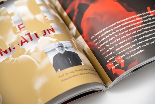 Nahaufnahme einer aufgeschlagenen Broschüre mit goldfarbenem und schwarzem Hintergrund. Links ein Porträtfoto eines Professors mit Text, rechts ein dynamisches Bild in Rot-Schwarz.