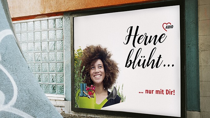 Eine Werbetafel zeigt eine fröhliche junge Frau mit lockigem Haar zwischen Wildblumen und der Aufschrift Herne blüht. 
