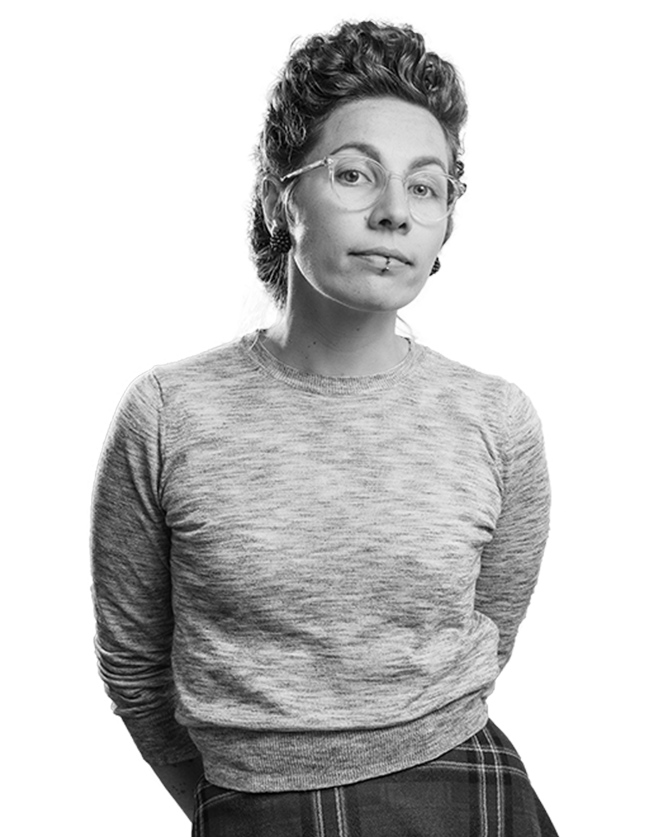 Frau in grauem Pullover und kariertem Rock, mit Brille und kurzen Haaren, schaut leicht lächelnd.