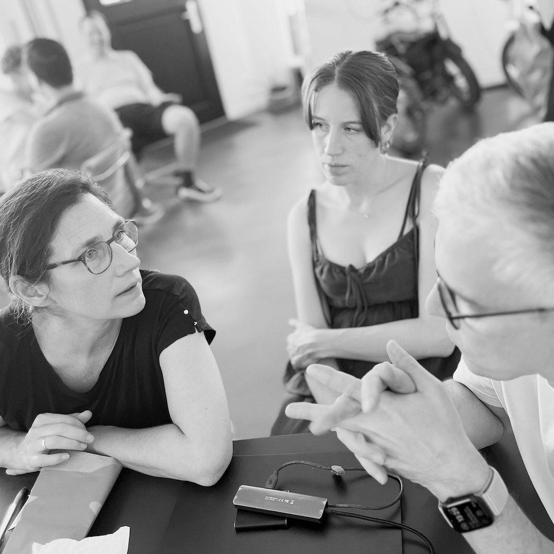 Drei Personen diskutieren an einem Tisch, im Hintergrund weitere unscharfe Figuren.