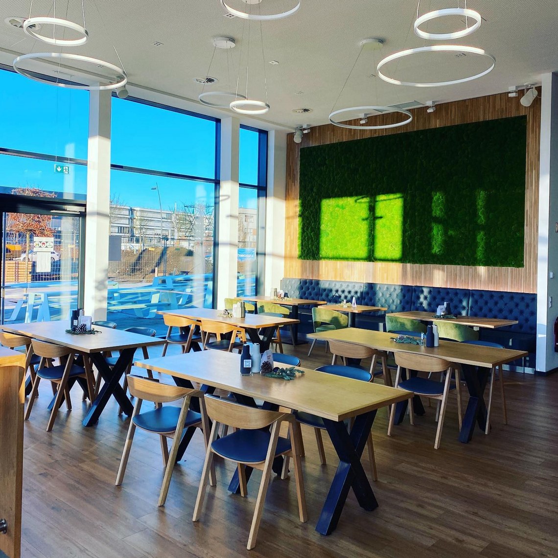 Helles Café-Interieur mit Holzmöbeln, grüner Mooswand und modernen Ringlampen.