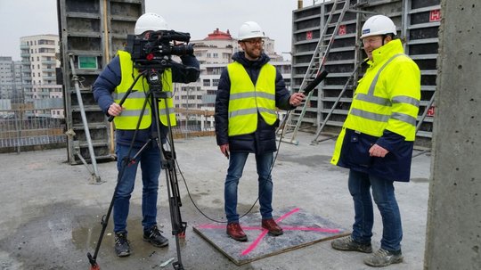 Drei Personen mit Schutzhelmen und Warnwesten auf einer Baustelle, eine bedient eine Kamera, die anderen führen ein Interview.