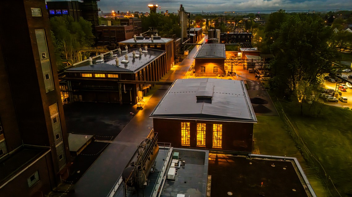 Industrielle Anlage bei Nacht, warm beleuchtete Gebäude und umgebende Bäume.