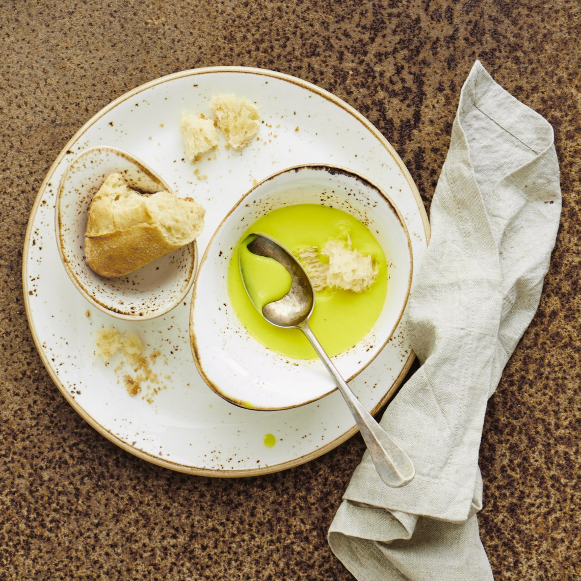 Teller mit Brot, Olivenöl und Löffel, daneben eine graue Serviette auf rustikalem Hintergrund.