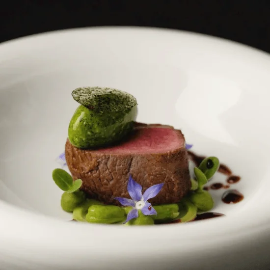 Edle Fleisch-Präsentation mit grünem Pesto, grünen Bohnen und lila Blüten auf weißem Teller.