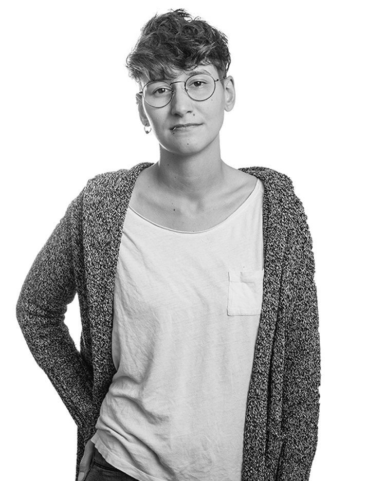 Frau mit Brille und lockerer Kleidung, neutraler Gesichtsausdruck vor weißem Hintergrund.