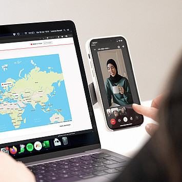 Neben einem offenen Laptop, dass eine Weltkarte zeigt, steht ein Handy. Auf dem Handy läuft eine Videokonferenz, bei der eine enstannte junge Frau mit Kopftuch und Kaffeetasse zu sehen ist.