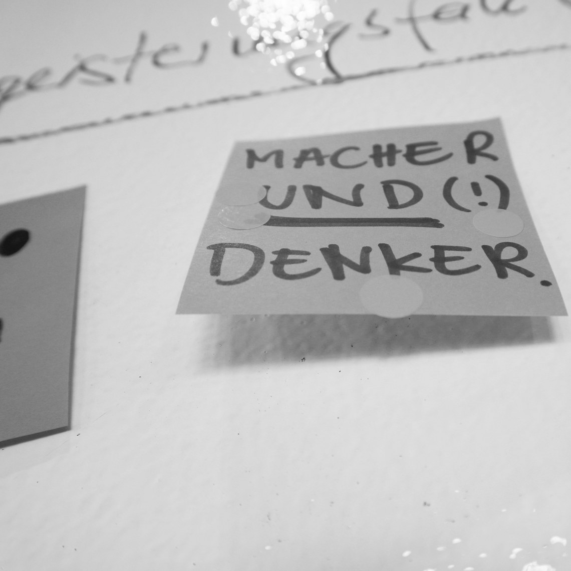 Notizzettel mit der Aufschrift "MACHER UND (!) DENKER" an einer Wand.