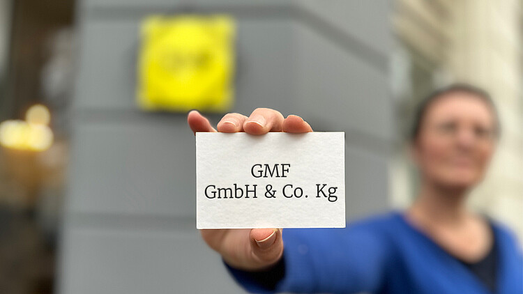 Mitarbeiterin hält Visitenkarte mit "GMF GmbH & Co. KG" Aufschrift vor Firmenschild