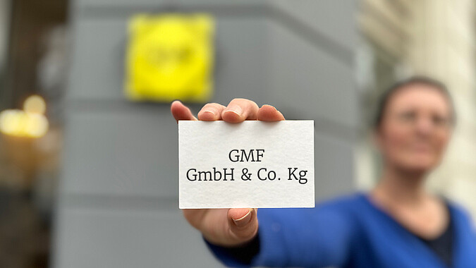 GMF GmbH & Co. KG Visitenkarte, gehalten von GMF-Mitarbeiterin vor Firmenschild