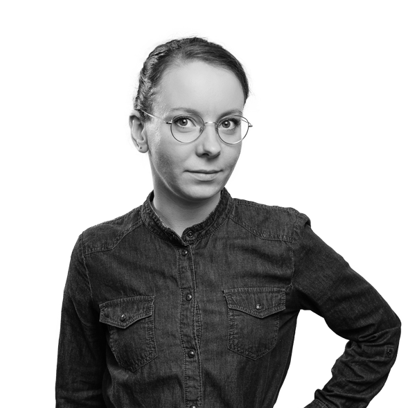 Frau mit Brille, Hand in die Hüfte gestützt, dunkles Hemd, vor schwarzem Hintergrund.