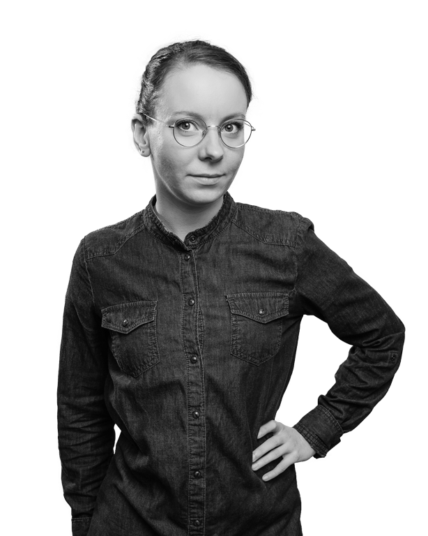 Frau mit Brille, Hand in die Hüfte gestützt, dunkles Hemd, vor schwarzem Hintergrund.
