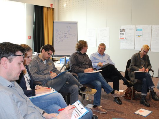 Eine Gruppe von Personen sitzt in einem Konferenzraum, einige schreiben auf Klemmbrettern. Im Hintergrund ist ein Flipchart und Plakate an der Wand zu sehen.