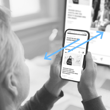 Person hält ein Smartphone, das auf einen PC-Bildschirm zeigt.